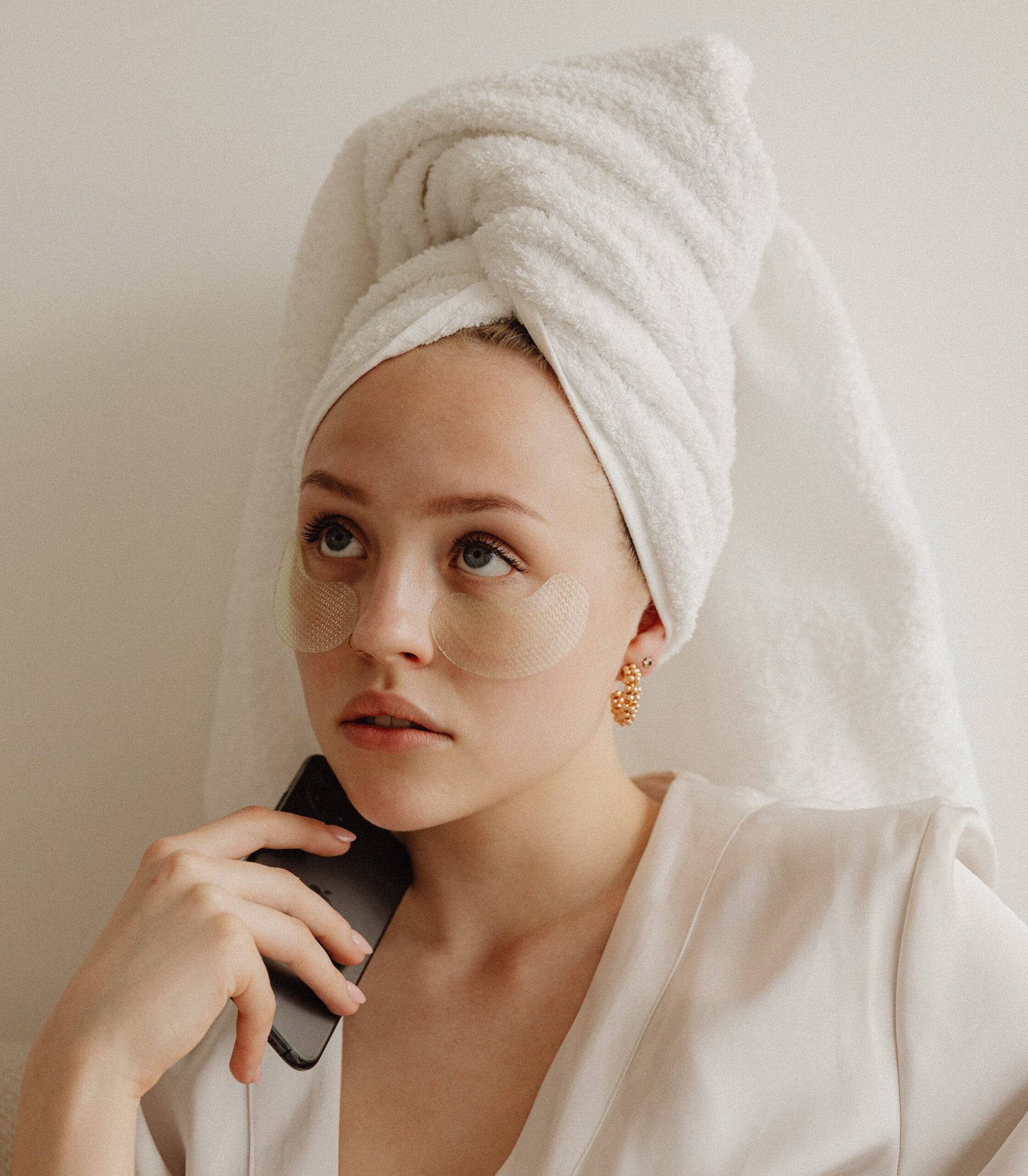 Femme en peignoir, une serviette enroulée autour de ses cheveux, regardant vers le haut avec un téléphone à la main et des patchs en dessous des yeux