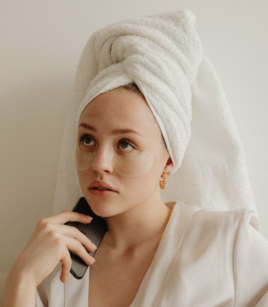 Femme en peignoir, une serviette enroulée autour de ses cheveux, regardant vers le haut avec un téléphone à la main et des patchs en dessous des yeux